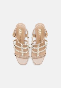 Sandalias de tiras beige con acentos de tachuelas doradas, acabado brillante y diseño de punta cuadrada; cuentan con una correa ajustable en el tobillo.