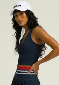 Top corto de canalé navy con escote en V, acompañado de una falda plisada navy con acentos de rayas rojas y blancas. Gorra de malla blanca con el logo "Wilson".