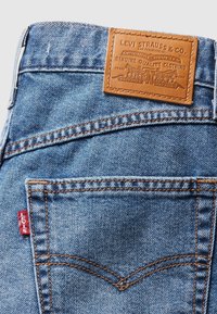 Jean Levi's en denim bleu montrant une poche arrière avec couture orange, étiquette rouge Levi's et patch en cuir beige.