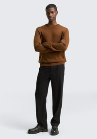 Brauner gestrickter Pullover mit Rundhalsausschnitt, strukturierter Stoff; kombiniert mit schwarzen Nadelstreifenhosen und schwarzen klobigen Stiefeln. Modell steht mit verschränkten Armen.