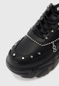 Sapatilha plataforma preta com um upper sintético liso, acentuado por tachas prateadas e detalhe em corrente. Apresenta atacadores pretos e uma sola texturizada.
