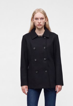 Jeune femme aux longs cheveux blonds portant un manteau noir croisé et un jean bleu, debout devant un fond clair uni.