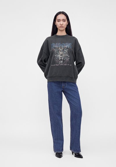 Femme portant un sweat-shirt oversize foncé avec un motif léopard et un texte bleu, associé à un jean droit bleu et des chaussures noires pointues.