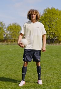 Nike spordirõivad, mis koosnevad heledast beežist tipust punase logoga ja mustadest lühikestest pükstest. Valge jalgpall hoitakse vasakus käes, taustaks muruväli.