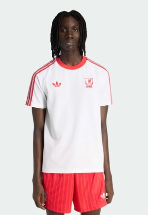 Jeune personne à la peau foncée et aux dreadlocks portant un maillot de football Adidas blanc et rouge avec un short assorti, debout devant un fond clair.