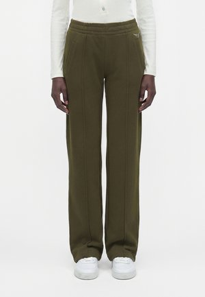 Personne portant un pantalon large vert olive avec des coutures devant, des poches latérales, un petit logo, des baskets blanches et un haut blanc à manches longues.