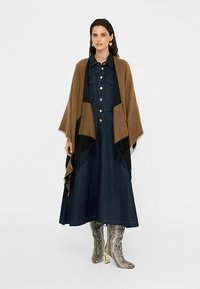 Barna sál fekete díszítéssel és rojttal, sötét farmerruha fölött, amely gombos elülső résszel és zsebekkel rendelkezik. Ezüst színű kígyómintás csizmák.