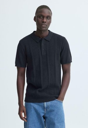 Zwart geribd poloshirt met een kraag en een knoopsluiting met drie knopen, gecombineerd met lichtblauwe spijkerbroek. Korte mouwen, gestructureerde stof.