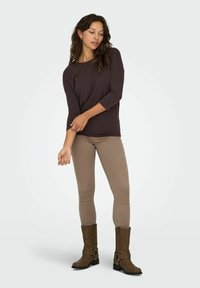 Top a maniche lunghe marrone, jeans aderenti marrone chiaro e stivali marroni al ginocchio con un dettaglio a fibbia. Completo semplice e casual con una texture liscia.