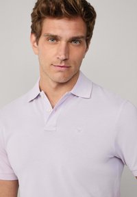 Světle fialová polo košile s malým vyšitým logem na hrudi, klasickým límečkem a tříknoflíkovou légou. Měkká textilie.