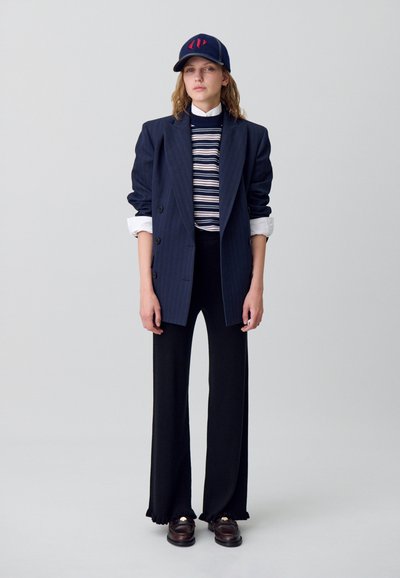Blazer oversize a rayas marinas, top de punto a rayas, pantalones anchos negros y zapatos oscuros, combinados con una gorra marina con un logo rojo.