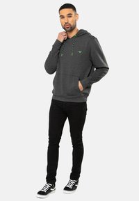 Sudadera gris con cordones verdes y pequeño logo, bolsillo frontal, puños y dobladillo acanalados; combinada con jeans skinny negros y zapatillas negras.