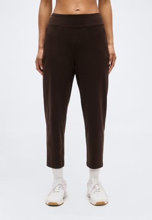 Μπροστινή όψη ατόμου που φοράει σκούρο καφέ κοντά παντελόνια, λευκές κάλτσες και λευκά αθλητικά παπούτσια, στέκεται σε απλό φόντο.