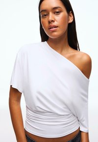 Top bianco con spalle scoperte in tessuto morbido con design drappeggiato; presenta una vita arricciata. Texture liscia; maniche corte e larghe.