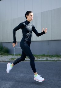 Sort langærmet atletisk top med teksturerede detaljer, parret med sorte leggings og hvide sneakers med neongrønne sokker. Løbeaction fanget.