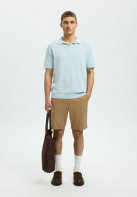 Svijetloplava pletena polo majica, kaki šortsevi, smeđe cipele, bijele čarape do članka i smeđa torba. Model stoji na jednoličnoj pozadini.