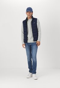 Gilet matelassé bleu marine sur un pull gris clair, associé à un jean bleu et des baskets blanches, coiffé d'une casquette bleu clair.