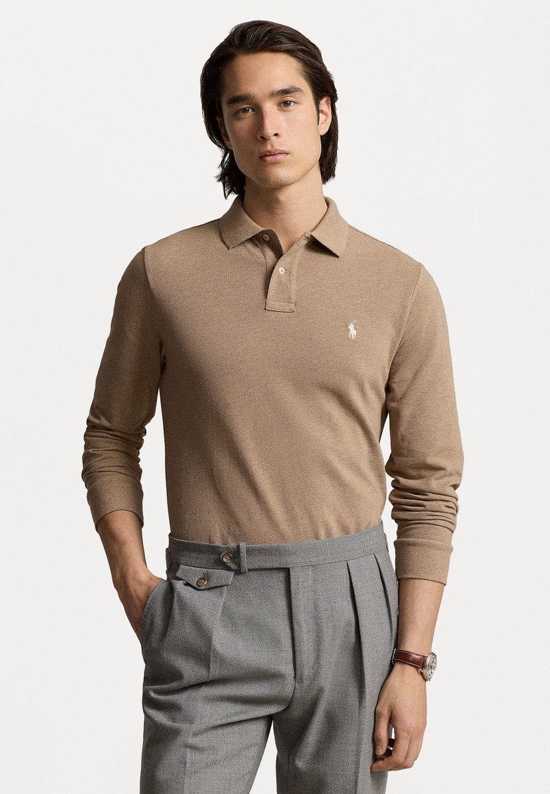 Beige polo met lange mouwen en een kraag, voorzien van een klein logo; gecombineerd met grijze geplooide broek met zijzakken en een riem.
