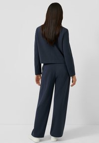 Granatowy dwuczęściowy strój z krótką marynarką i spodniami o szerokich nogawkach. Teksturowana tkanina i czyste linie, zestawione z białymi trampkami.
