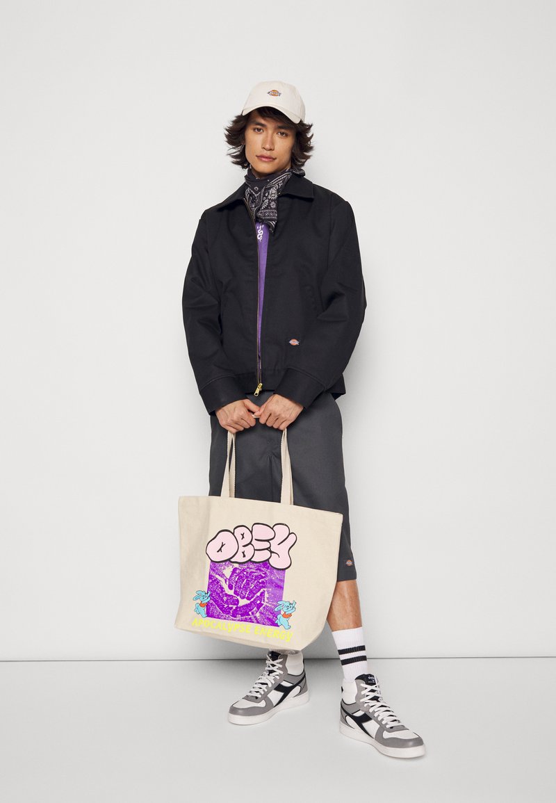 Chaqueta negra con cremallera, forro morado, gorra de béisbol color crema, bandana, pantalones cortos grises, bolsa beige con gráfico colorido, zapatillas grises y blancas con rayas negras.