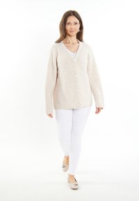 Cardigan beige in maglia con scollo a V, bottoni frontali e maniche lunghe, abbinato a pantaloni bianchi e scarpe chiare. Tessuto morbido e vestibilità rilassata.