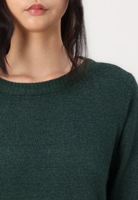 Femme aux cheveux raides et foncés portant un pull en maille vert foncé à encolure ronde, sur un fond neutre.