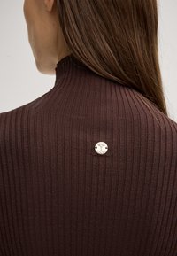 Gerippter brauner Rollkragenpullover mit hohem Kragen, der einen goldfarbenen Logo-Knopfakzent auf der Rückseite hat. Breiten- und Texturdetails sind sichtbar.