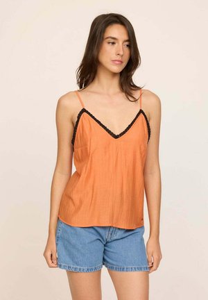 Camisole orange sans manches avec un bord en dentelle noire, présentant un tissu texturé subtil et un décolleté en V. Associée à un short en denim.