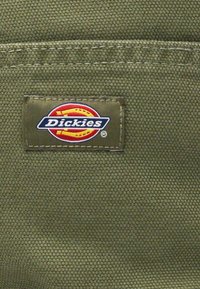 Primeiro plano de tecido verde-oliva com um rótulo retangular da marca Dickies, costurado, apresentando um logótipo em vermelho, amarelo, branco e azul-marinho.