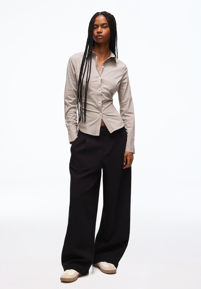 Femme avec de longues tresses porte une chemise à carreaux beige boutonnée, un pantalon large noir et des baskets blanches, debout avec une main dans la poche.