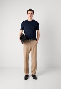 Tamsiai mėlynas trumpomis rankovėmis marškinėliai su subtilia tekstūra, smėlio spalvos kelnės, juodas diržas ir blizgantys juodi batai, laikantis juodą rankinę.