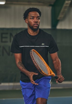 Camisola desportiva preta com risca branca e logótipo da Lacoste, acompanhada de calções azuis. Asegura uma raquete de ténis laranja, com um fundo de um campo desportivo.