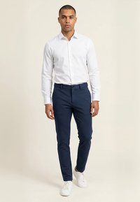 Mand stående iført en hvid langærmet skjorte, marineblå smalle bukser og hvide sneakers mod en ensfarvet baggrund.