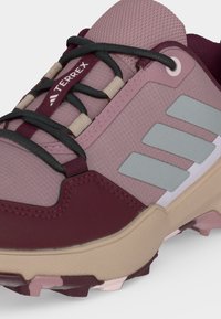 Gros plan d'une chaussure de trail Adidas Terrex violette et beige avec des lacets noirs, trois bandes gris clair et une semelle robuste.