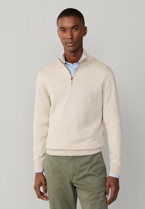 Man draagt een beige sweater met kwartritssluiting over een lichtblauw overhemd en olijfgroene broek, staand tegen een effen grijze achtergrond.