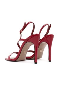 Des sandales à talons hauts en cuir verni rouge présentent un design à lanières, une boucle à la cheville réglable et une semelle intérieure beige contrastante avec un détail de logo.