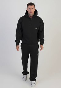 Svart zip-up hoodie med en känguruficka, kombinerad med matchande svarta joggers. Mjukt tyg, minimal vit märkning på båda plaggen.