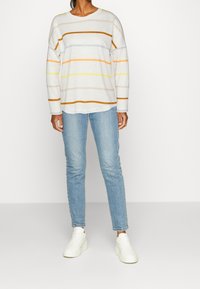 Weißes Langarmhemd mit horizontalen Streifen in Orange, Gelb und Beige, kombiniert mit hellblauen Jeans und weißen Sneakers.