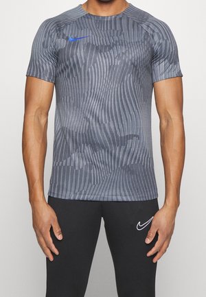 Homme portant un t-shirt Nike gris texturé à manches courtes et un pantalon Nike noir, debout avec les bras détendus le long du corps sur un fond uni.