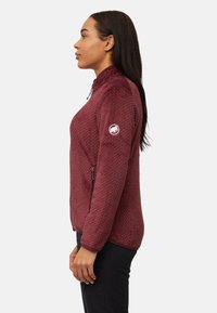 Giacca in pile color bordeaux con motivo a rilievo, colletto alto, tasche con cerniera e logo sul braccio superiore. Superficie liscia, design aderente.