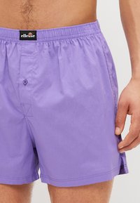 Λεβάντα μαγιό φτιαγμένα από ελαφρύ ύφασμα με ελαστική ζώνη μέσης, διαθέτοντας κλείσιμο με κουμπί, ραμμένα διακοσμητικά στοιχεία και πλάγιες σχισμές.