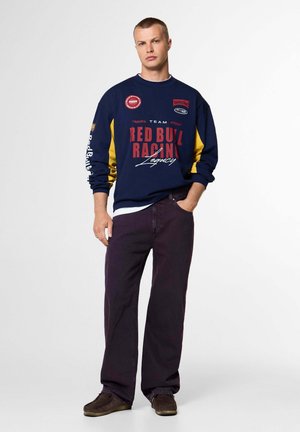 Junger Mann, der einen marineblauen und gelben Red Bull Racing Sweatshirt, dunkle lockere Hose und braune Schuhe trägt, posiert mit einer Hand in der Tasche.