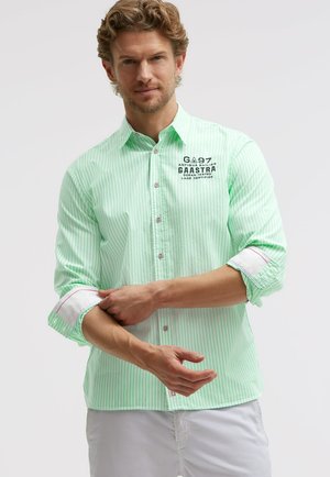 Mann krempelt die Ärmel eines hellgrün-weiß gestreiften Hemdes mit bedrucktem Schriftzug auf der Brust hoch, trägt weiße Hose.