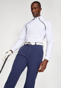 Weißes Langarm-Golfschirt mit Reißverschlusskragen, das mit schwarzem Piping verziert ist. Kombiniert mit dunkelblauen Hosen und einem weißen Gürtel, hält einen Golfschläger.