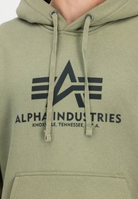 Svetlozelená mikina s kapucňou s čiernym logom Alpha Industries, textom "Knoxville, Tennessee, U.S.A." a šnúrkami cez náprsné vrecko.