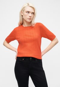 Νεαρή γυναίκα με ξανθά μαλλιά, φοράει πορτοκαλί πουλόβερ με κοντά μανίκια και μαύρο τζιν, στέκεται με τα χέρια στη μέση, κοιτάζοντας πλάγια.