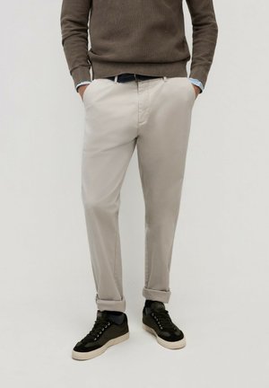 Hombre vestido con pantalones beige remangados, suéter marrón sobre camisa azul y zapatillas negras, de pie con las manos en los bolsillos sobre un fondo liso.