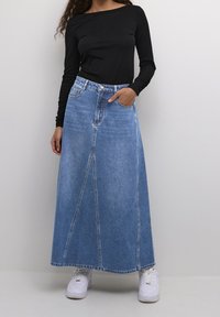Falda larga de mezclilla en azul claro, con cintura alta, bolsillos frontales y detalles de costura vertical. Combinada con una camiseta de manga larga negra y zapatillas blancas.