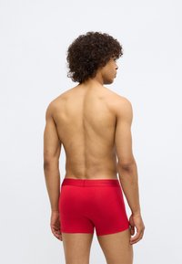 Boxer briefs vermelhos com uma textura suave e um ajuste justo. Apresenta uma cinta com a marca Calvin Klein. Vista traseira, modelo está sem camisa.