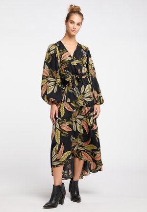 Vrouw die een lange zwarte jurk draagt met roze en groene bladmoten, pofmouwen, een gestrikte taille en zwarte enkellaarsjes, staand tegen een witte achtergrond.